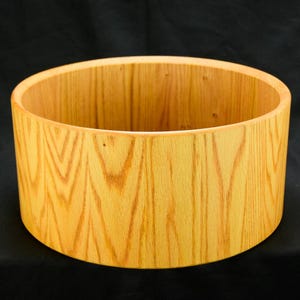 May include: A cylindrical wooden bowl with a light brown finish. The bowl has a smooth, rounded rim and a natural wood grain pattern. The interior and exterior are the same color. The bowl is likely handmade.