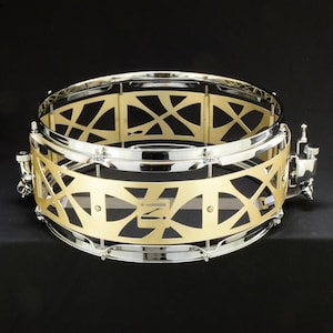 May include: A cylindrical snare drum shell featuring a polished chrome frame and a decorative gold-colored metal wrap with a geometric cutout pattern. The drum includes a side-mounted tension adjustment mechanism and a horizontal wire snare strainer.