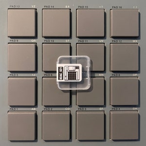 Peut inclure: Un pad de production musicale électronique gris et noir avec 16 boutons étiquetés avec des lettres et des symboles. Un boîtier en plastique transparent avec un autocollant noir et blanc qui dit "BDAC_SD1" est sur le dessus du pad.