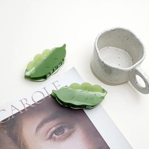 May include: Two green pea pod-shaped hair clips and a speckled white ceramic mug with a handle. The clips are a translucent green with a scalloped edge, and the mug has a rounded shape.