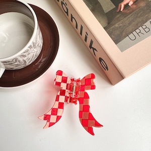 May include: A red and pink checkered bow-shaped hair clip. The clip is made of translucent plastic and has a metal hinge. A white teacup and saucer with a floral design are in the background, along with a book.