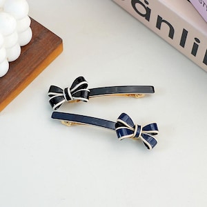 Peut inclure: Deux barrettes à cheveux avec des nœuds. L'une est bleu marine avec un nœud blanc, l'autre est noire avec un nœud blanc. Les barrettes ont un fermoir doré.