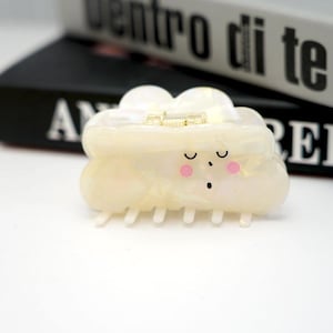 Op de afbeelding: Een witte wolkvormige haarspeld met een schattig slapend gezicht en roze wangen. De speld heeft een gouden sluiting en een rij tanden aan de onderkant.