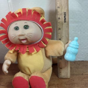 May include: A plush doll dressed as a lion, featuring a red and yellow mane, a yellow body, and red feet. The doll has green eyes and is holding a blue baby bottle. A wooden ruler is next to the doll.