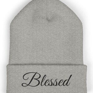 May include: A grey beanie hat with the word "Blessed" in black script. The hat is made of a soft, knitted material and has a folded brim. The beanie is a neutral colour and suitable for all.