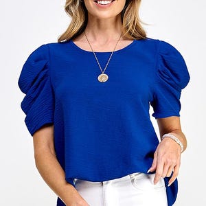 May include: A royal blue short-sleeved top with puffed sleeves and a round neckline. The top is made of a textured fabric and has a slightly longer back hem. The model is wearing a gold necklace with a round pendant.