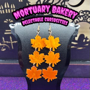 May include: A pair of orange maple leaf dangle earrings. Each earring has three leaves, with the top leaf being the largest and the bottom leaf being the smallest. The leaves are made of a translucent orange material with a marbled effect. The earrings are hanging from gold-colored hooks.