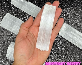 May include: Four white selenite sticks, also known as selenite wands, are shown in the image. The sticks are smooth and have a striated texture. The sticks are held in a person's hand.
