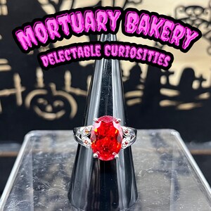 May include: A silver ring with a large, oval-shaped, red gemstone. The ring is set on a black ring holder. The background is a black and white image of a spooky scene with the text "Mortuary Bakery Delectable Curiosities".