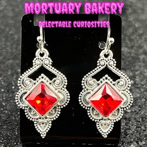 May include: A pair of silver dangle earrings with red crystal accents. The earrings feature a delicate, ornate design with a square-shaped crystal in the center.