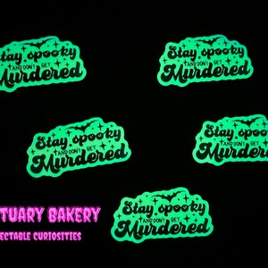 Peut inclure: Cinq autocollants phosphorescents avec le texte "Stay spooky and don't get murdered" dans une police rétro. Les autocollants sont verts et noirs et ont un motif de chauve-souris et d'étoiles. Le texte "Mortuary Bakery Delectable Curiosities" est également visible en rose.