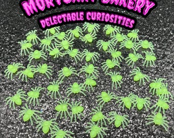 May include: A collection of 30 glow-in-the-dark plastic spiders. The spiders are green and have eight legs. The spiders are arranged in a random pattern on a sparkly black surface. The text "Mortuary Bakery Delectable Curiosities" is written in pink letters at the top of the image.