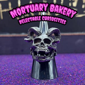 May include: A silver skull ring with horns, displayed on a black stand. The ring features a detailed skull with fangs and horns. The text "Mortuary Bakery Delectable Curiosities" is visible in the background.