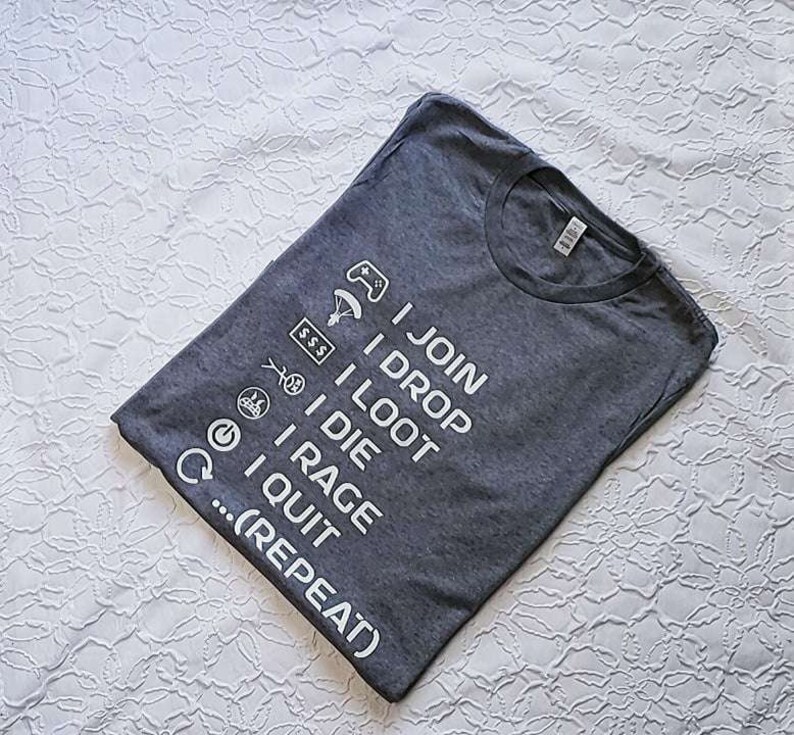May include: Charcoal grey t-shirt featuring white text and gaming symbols. The text on the shirt says "I JOIN, I DROP, I LOOT, I DIE, I RAGE, I QUIT... (REPEAT)". The t-shirt is folded on a white, patterned surface.