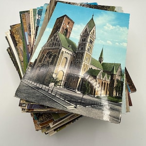 May include: A stack of vintage postcards featuring a variety of colorful printed images. The top postcard displays a large stone cathedral with green roofs and tall towers against a light blue sky, with a paved plaza in the foreground.