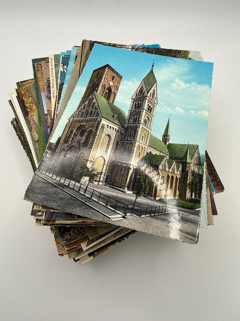 May include: A stack of vintage postcards featuring a variety of colorful printed images. The top postcard displays a large stone cathedral with green roofs and tall towers against a light blue sky, with a paved plaza in the foreground.