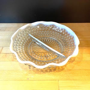 Puede incluir: Un plato de servir dividido de vidrio transparente con borde festoneado y un utensilio de servir plateado. El plato tiene un patrón texturizado de pequeños círculos en la sección exterior y un diseño de rayos de sol en el centro.