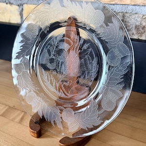 Puede incluir: Plato de vidrio transparente con diseño grabado de cardos y hojas. El plato se exhibe sobre un soporte de madera, mostrando el intrincado patrón botánico. El diseño del plato presenta hojas y cabezas de flores detalladas.
