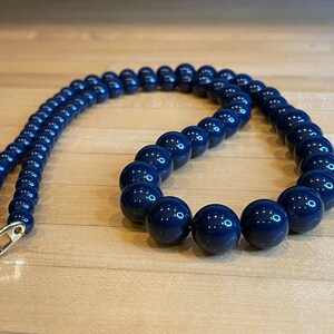 Puede incluir: Un collar de cuentas azul marino con un cierre dorado. El collar presenta cuentas grandes y redondas que están espaciadas uniformemente. Las cuentas tienen un acabado brillante y reflejan la luz. El collar se exhibe sobre una superficie de madera clara.