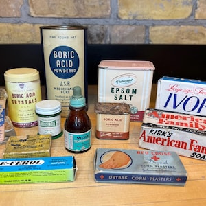 May include: A collection of vintage apothecary items, including Boric Acid powder and crystals, Epsom Salt, Ivory soap, and various medicinal products. The items are arranged on a wooden surface against a brick background.