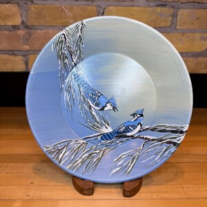 May include: Hand-painted decorative plate with two blue jays on a branch with white and green leaves. The plate has a light blue background and is displayed on a wooden stand. The artwork is detailed and vibrant.