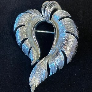 Puede incluir: Broche de tono plateado con un diseño de plumas en forma de corazón.