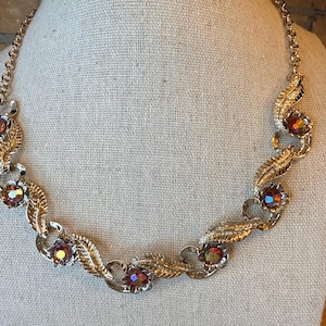 May include: A gold-toned necklace with a floral and leaf design. The necklace features iridescent, round gemstones set within the floral elements. The leaves and chain have a textured appearance, creating a vintage aesthetic.