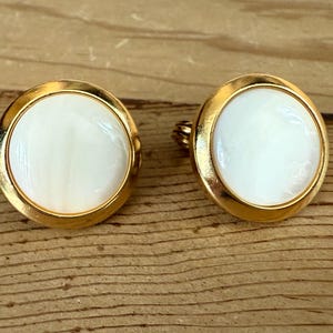 Könnte beinhalten: Ein Paar goldfarbene Manschettenknöpfe mit einem runden, weißen, perlmuttartigen Mittelteil. Die Manschettenknöpfe sind vor einem Holzgrund platziert, was ihre polierte Oberfläche und ihr klassisches Design hervorhebt. Geeignet für formelle Kleidung.