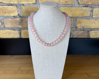 May include: A pink beaded necklace displayed on a white mannequin bust. The necklace is composed of round, light pink beads. The background features a brick wall and a wooden surface.
