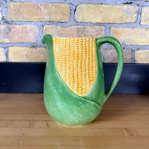 May include: A ceramic pitcher shaped like an ear of corn. The pitcher has a yellow corn cob design in the center, wrapped in green husks. It has a green handle and spout, and sits on a wooden surface.