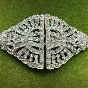 Puede incluir: Un broche para ropa de metal plateado con forma de diamante alargado y simétrico. La superficie presenta detalles intrincados con patrones de filigrana calados y está incrustada con numerosos diamantes de imitación redondos y transparentes.