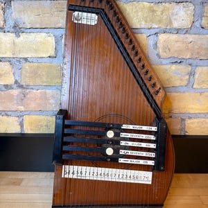 Può includere: Un'autoharp in legno vintage di forma trapezoidale. Lo strumento ha numerose corde, piroli e una serie di barre etichettate. Il corpo è di colore marrone caldo, con accenti neri ed etichette bianche. L'autoharp è esposta contro un muro di mattoni.