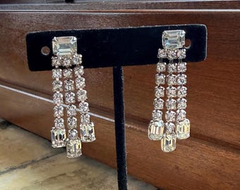 May include: A pair of silver-toned dangle earrings with clear, sparkling rhinestones. Each earring features a large, square-cut stone at the top, with multiple strands of smaller stones cascading downwards. The earrings are displayed on a black stand.
