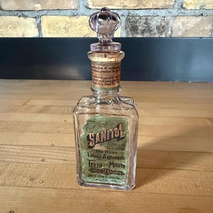 May include: A vintage glass bottle with a decorative stopper. The bottle has a label that reads "Sanitol" and "Liquid Antiseptic for Teeth and Mouth." The stopper is a light purple color. The bottle is rectangular and sits on a wooden surface.