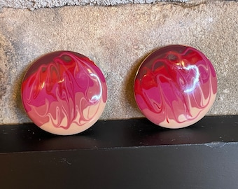 May include: A pair of round, pink and red swirl patterned earrings. The earrings are made of a smooth, shiny material.