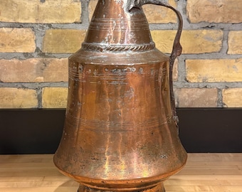 May include: A large, antique copper pitcher with a lid and handle. The pitcher has a bulbous base and a tapered body, with decorative bands and a textured surface. The copper has a warm, reddish-brown patina, suggesting age and use. The pitcher is displayed on a wooden surface.