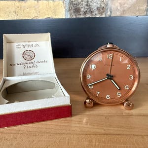 Peut inclure: Un réveil CYMA de couleur cuivre avec un cadran rond et une boîte assortie. L'horloge a un cadre de couleur cuivre, un cadran blanc avec des chiffres noirs et trois pieds sphériques. La boîte est blanche avec une bordure rouge et le logo CYMA.