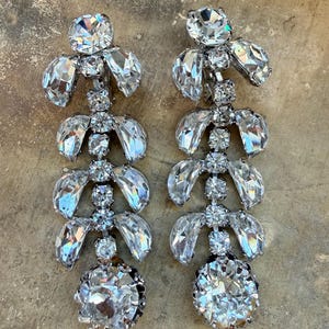 May include: A pair of dazzling, silver-toned chandelier earrings. Each earring features multiple tiers of clear, faceted crystals in various shapes, culminating in a large, round crystal at the bottom. The earrings are designed to catch and reflect light.