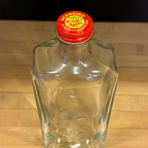 以下が含まれることがあります： 赤いキャップが付いた透明なガラス瓶。キャップには「Dickinson's Double Distilled Witch Hazel」と書かれています。ボトルは独特の角張った形状で、木製の表面に置かれています。