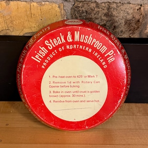 Könnte beinhalten: Eine runde, rote Dose Irish Steak & Mushroom Pie with Puff Pastry. Das Etikett enthält Backanweisungen und den Text "PRODUCT OF NORTHERN IRELAND". Die Dose steht auf einer Holzoberfläche.