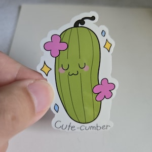 May include: A sticker featuring a cartoon cucumber with a smiling face, pink flowers, and sparkling accents. The text "Cute-cumber" is printed at the bottom. The cucumber is green with vertical lines.