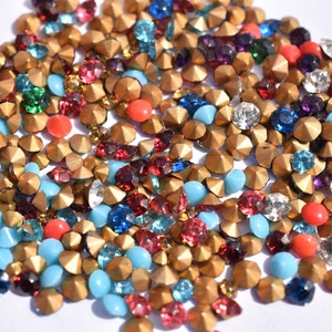 May include: A large assortment of colourful rhinestones and gold-toned studs. The rhinestones are in various colours, including red, blue, green, turquoise, and clear. The studs are all gold-toned and have a pointed top.