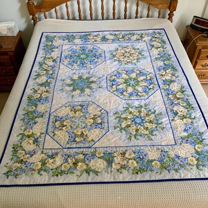 May include: A rectangular quilt featuring a white background with a blue floral border and four large octagonal panels. Each panel displays a symmetrical arrangement of blue and white flowers and green foliage. The quilt is finished with a blue binding.
