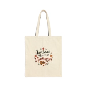 May include: A beige canvas tote bag featuring a central graphic with the text "Viviendo Nuestras Tradiciones" in dark brown and red script. The design includes small orange and red flowers, green leaves, tiny hearts, and a small brown guitar.