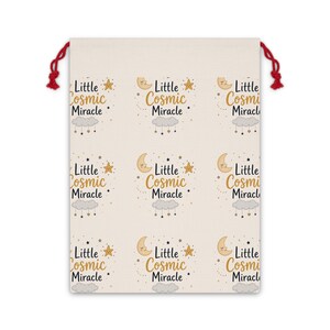 Peut inclure: Un sac à cordon de couleur crème avec un motif répétitif du texte Little Cosmic Miracle en écriture noire et dorée. Le design comprend des illustrations d'un croissant de lune endormi, d'une étoile et d'un nuage gris avec des étoiles suspendues.