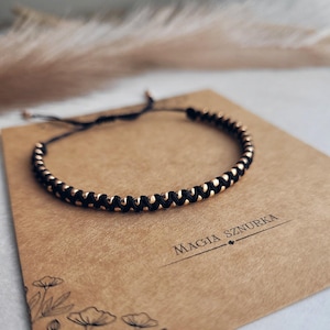 Può includere: Un braccialetto con perline nere e dorate, con un cordoncino nero e una chiusura a nodo scorrevole. Il braccialetto è su uno sfondo marrone con il testo "Magia Sznurka".