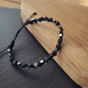May include: A black and silver beaded bracelet with a black cord closure. The bracelet features black lava rock beads and silver square beads.
