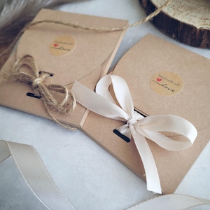 May include: Two brown paper packages tied with twine and cream-colored ribbon. Each package has a small round sticker with the words "handmade with love" and a heart. The packages are on a white surface.