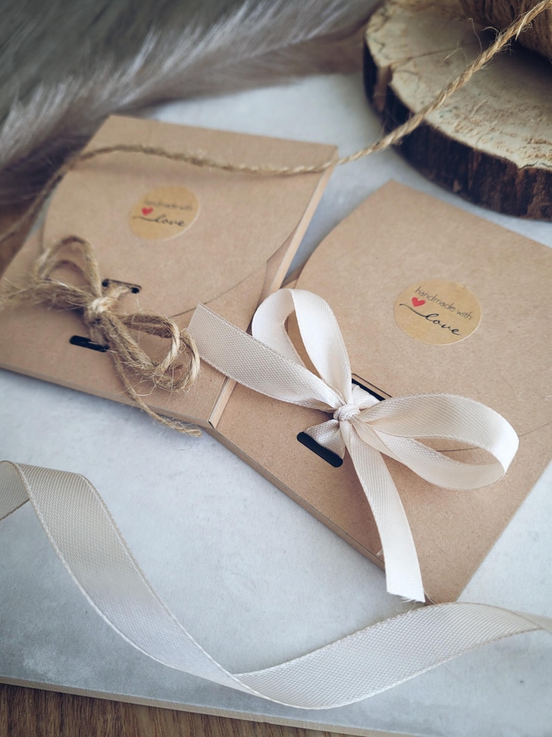 May include: Two brown paper packages tied with twine and cream-colored ribbon. Each package has a small round sticker with the words "handmade with love" and a heart. The packages are on a white surface.