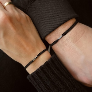 Puede incluir: Dos pulseras de cordón trenzado negro con detalles metálicos plateados. Las pulseras se llevan en las muñecas, lo que sugiere un conjunto a juego o joyería para parejas. La imagen es un primer plano, destacando la textura y el diseño.